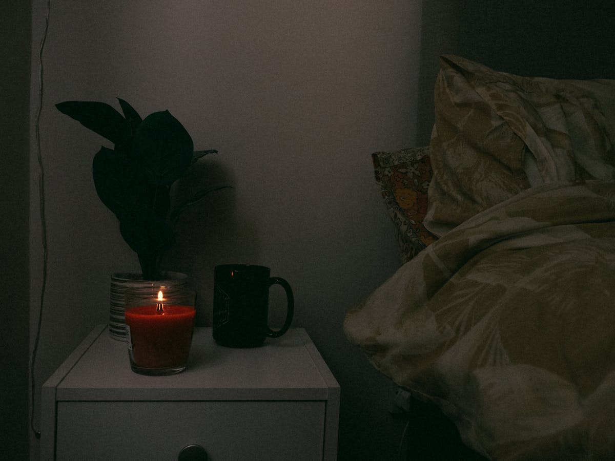 A cozy bedroom nightstand with a candle, warm mug, and small houseplant creating a peaceful low-light atmosphere at night